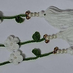 May include: Three crocheted green vines featuring white flowers with small central beads. Each vine ends with a cluster of white yarn tassels, accented by a sequence of small white pearls and metallic gold-toned beads near the tassel base.