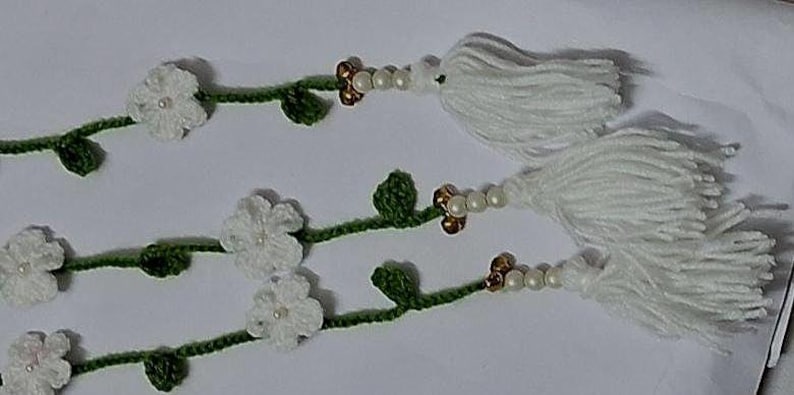 May include: Three crocheted green vines featuring white flowers with small central beads. Each vine ends with a cluster of white yarn tassels, accented by a sequence of small white pearls and metallic gold-toned beads near the tassel base.