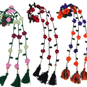 Könnte beinhalten: Drei lange, gehäkelte rankenartige Accessoires mit kleinen, bunten Blumen an grünen oder schwarzen Stielen. Jeder Strang endet mit einem dekorativen Perlenakzent und einer dicken, hängenden Garnquaste in passenden oder kontrastierenden Farben.