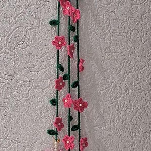 May include: A vertical hanging decoration featuring three long dark green crocheted vines adorned with small pink flowers and white beads. The bottom of the arrangement showcases two larger, multi-layered pink crocheted flowers with white centers.