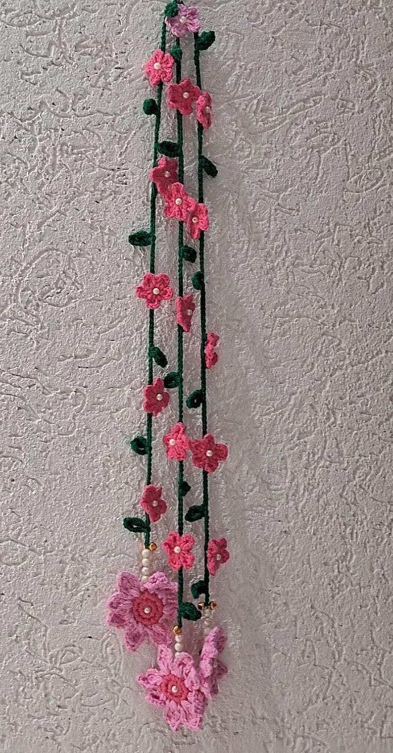 May include: A vertical hanging decoration featuring three long dark green crocheted vines adorned with small pink flowers and white beads. The bottom of the arrangement showcases two larger, multi-layered pink crocheted flowers with white centers.