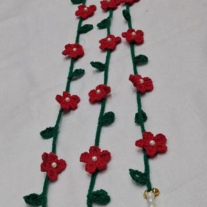 May include: Three vertical crochet garlands featuring green vines with small red five-petaled flowers, each centered with a white bead. The bottom of each vine terminates in a crocheted red strawberry with white dots, accented by white beads and gold bells.