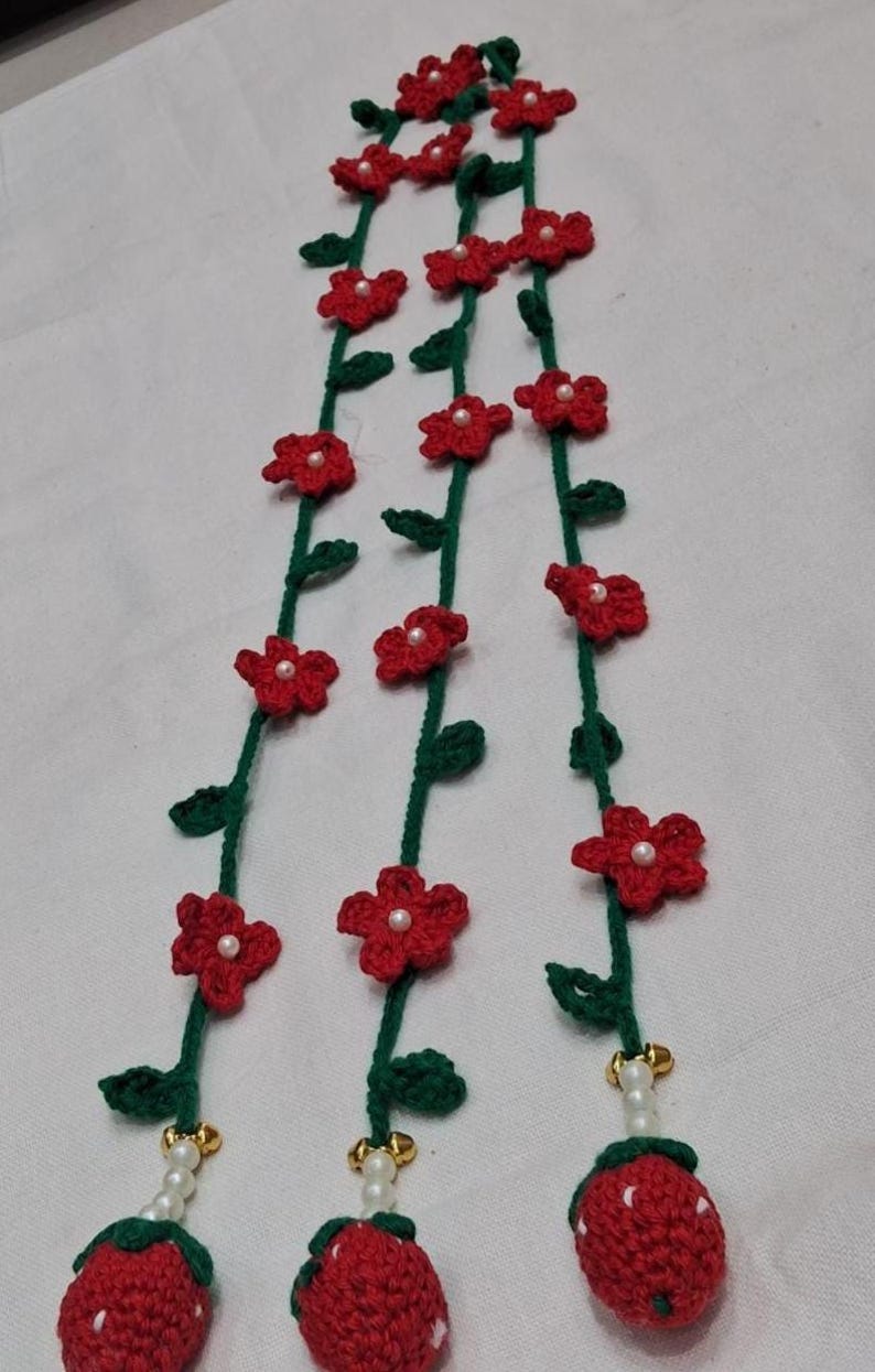 May include: Three vertical crochet garlands featuring green vines with small red five-petaled flowers, each centered with a white bead. The bottom of each vine terminates in a crocheted red strawberry with white dots, accented by white beads and gold bells.