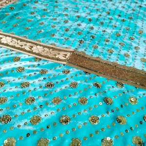 Op de afbeelding: Een turquoise blauwe stof met gouden pailletten in een bloemenpatroon. De stof heeft een gouden bies met een bloemenontwerp.