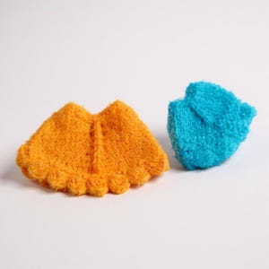 May include: Two small, handmade knitted items. One is orange, shaped like a cape with a scalloped edge. The other is a small, blue, textured item. Both are on a white background.