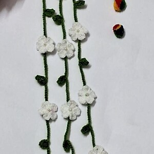May include: A crochet garland featuring three long green vines adorned with small white flowers, each centered with a tiny white bead. The vines terminate in white yarn tassels and decorative beads, accompanied by two small, round, multi-colored studs.