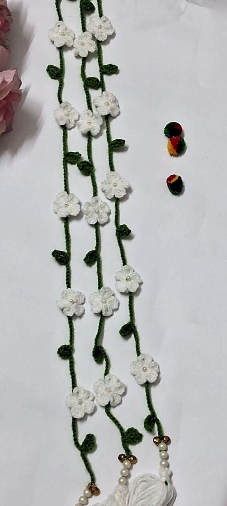 May include: A crochet garland featuring three long green vines adorned with small white flowers, each centered with a tiny white bead. The vines terminate in white yarn tassels and decorative beads, accompanied by two small, round, multi-colored studs.
