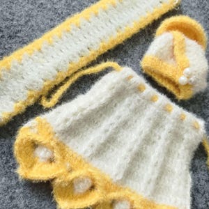 May include: A white and yellow knitted baby outfit with a headband, skirt, and booties. The skirt has a scalloped edge with yellow trim and white pom-poms.