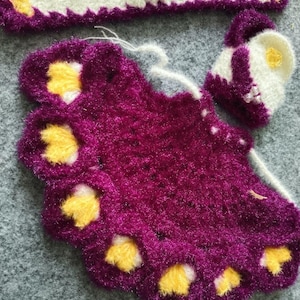 May include: A purple crocheted baby dress with yellow flower accents. The dress is made with a fuzzy yarn and has a white crocheted headband.