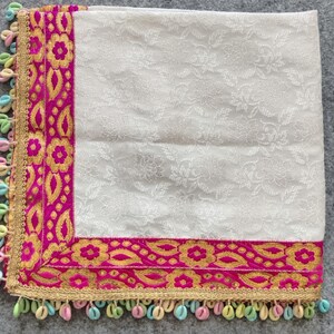 May include: White fabric square with a pink and gold embroidered border. The border has a floral pattern. The fabric is decorated with a white floral pattern. The square is trimmed with a row of colourful beads.