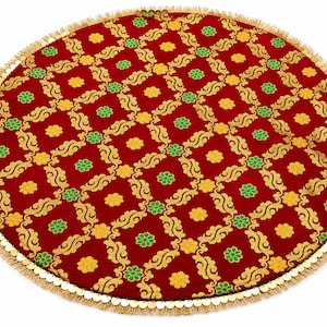 May include: A circular red fabric mat featuring a repeating gold scrollwork pattern interspersed with small yellow and green floral motifs. The edge of the mat is finished with a decorative gold fringe trim consisting of small metallic discs.