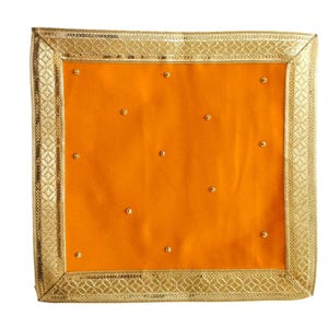 May include: A square, orange textile with gold beaded accents. The border is a gold, patterned trim with a sequined edge. The textile is likely used for decorative purposes.