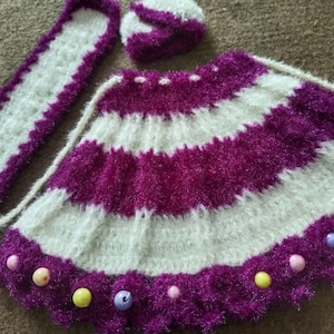 May include: A white and purple knitted baby dress with a matching headband and a white knitted shawl. The dress has a scalloped hem and is decorated with colourful beads.