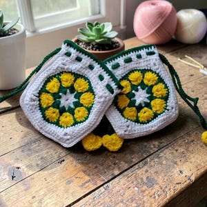 May include: Two white crochet bags with yellow and green floral designs. The bags have green drawstrings and yellow pom-poms. The bags are on a wooden surface, with succulents and yarn balls in the background.