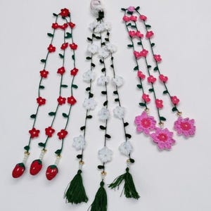 May include: Three long, vertical crochet garlands featuring thin green vines with small leaves. The left garland has red flowers and three red strawberry charms. The center has white flowers and green tassels. The right has pink flowers.