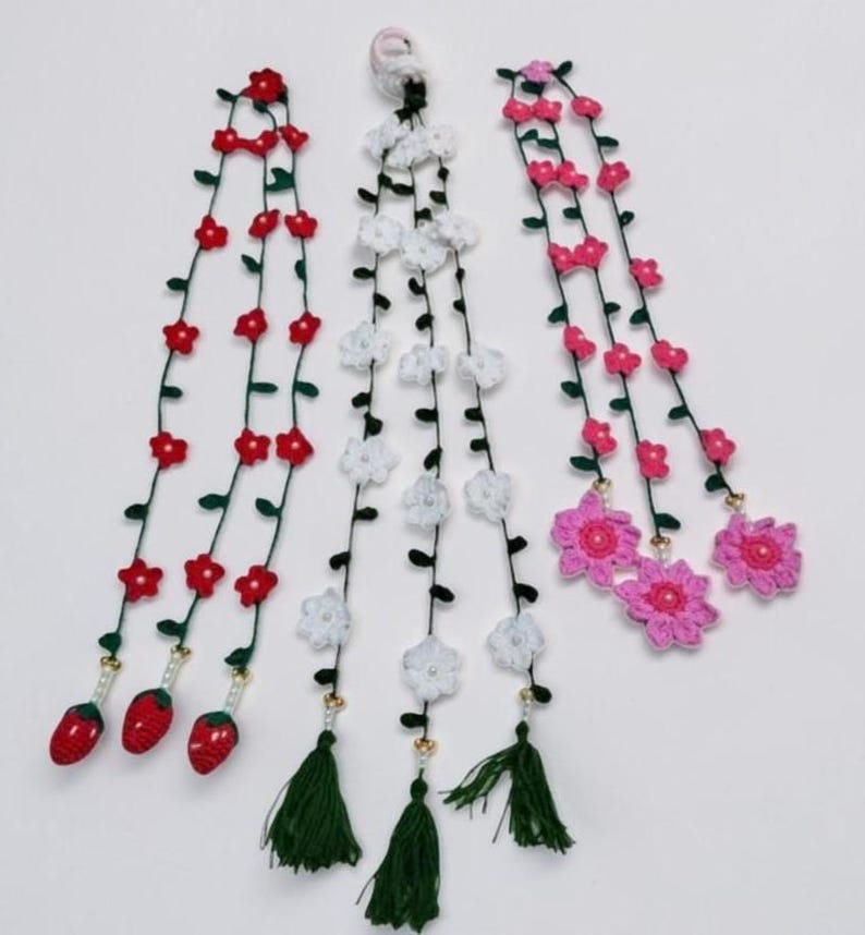 May include: Three long, vertical crochet garlands featuring thin green vines with small leaves. The left garland has red flowers and three red strawberry charms. The center has white flowers and green tassels. The right has pink flowers.