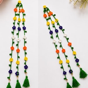 May include: Three long, green crocheted vine strands decorated with small, multi-colored flowers in yellow, orange, white, and purple. Each vine features tiny green leaves and ends in a green yarn tassel accented with small white and gold beads.