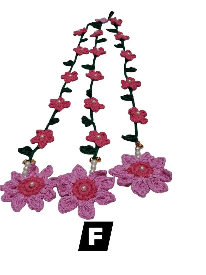 May include: A crochet garland featuring three large pink flowers with dark pink centers and white pearl accents, connected by green vines adorned with smaller pink flowers. The letter F is displayed in a black square at the bottom of the image.