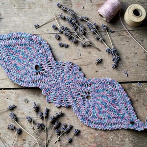 Peut inclure: Nappe en crochet faite à la main, aux tons de bleu, rose et violet, avec un motif ajouré décoratif. La nappe est posée sur une surface en bois rustique, avec des brins de lavande et des bobines de fil.