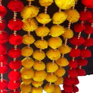 May include: A decorative garland featuring multiple vertical strands of vibrant red and bright yellow pom-poms. Each pom-pom is separated by small, shiny gold-colored beads strung along thin, flexible cords, creating a dense, colorful hanging display.