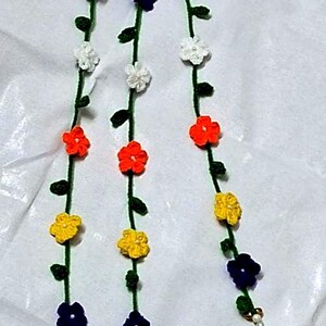 May include: A decorative hanging garland featuring three vertical green yarn strands adorned with small crocheted flowers in orange, yellow, white, and dark blue. Each strand includes tiny green leaves and terminates in a green yarn tassel.
