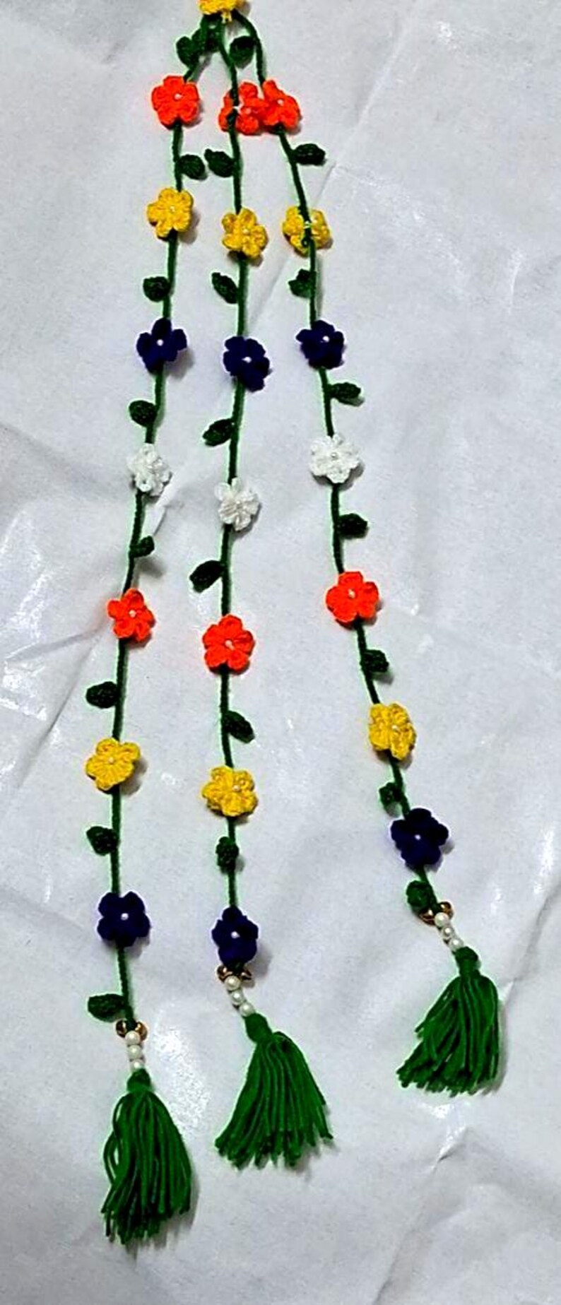 May include: A decorative hanging garland featuring three vertical green yarn strands adorned with small crocheted flowers in orange, yellow, white, and dark blue. Each strand includes tiny green leaves and terminates in a green yarn tassel.