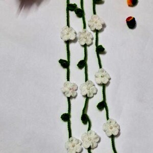 May include: A long, crocheted garland featuring green vines adorned with small white flowers, each centered with a tiny pearl bead. The garland ends in three white yarn tassels accented with gold-toned beads and additional pearl beads.