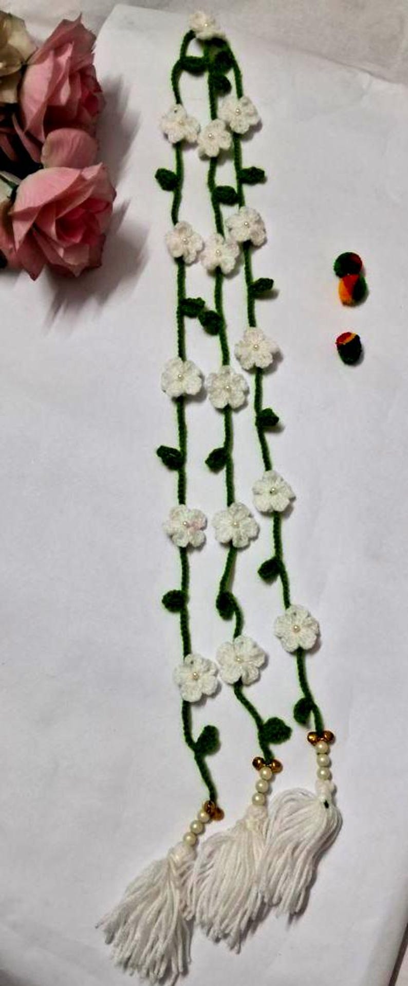 May include: A long, crocheted garland featuring green vines adorned with small white flowers, each centered with a tiny pearl bead. The garland ends in three white yarn tassels accented with gold-toned beads and additional pearl beads.