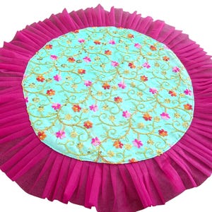 Pode incluir: Um tapete decorativo circular com um painel central de tecido azul-claro adornado com vinhas bordadas a ouro e pequenos detalhes florais cor-de-rosa e laranja, rodeado por uma larga borda de folhos de tule magenta vibrante.