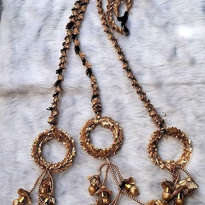 May include: Gold-colored decorative garland with three circular wreath-like elements. Each wreath has a tassel of gold-colored beads and bell-shaped ornaments. The garland is accented with black and gold detailing and a red rose.