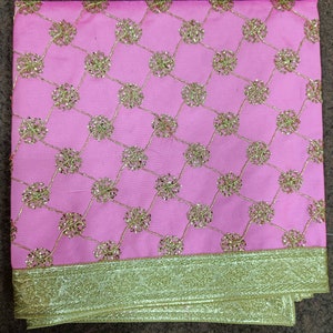 May include: Pink fabric with a gold lattice pattern and gold floral accents. The bottom edge features a wide gold border. This textile is suitable for various craft and design projects.