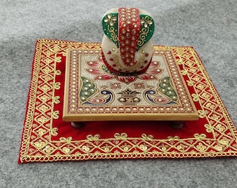 Può includere: Una statua di Ganesha in marmo bianco con accenti rossi, verdi e dorati si trova su una piattaforma quadrata in legno con una base in velluto rosso. La piattaforma è decorata con un motivo floreale e pavoni. La base ha un bordo ricamato in oro.