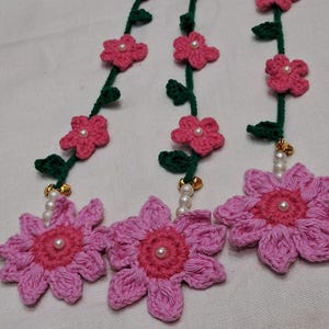 May include: Three crocheted floral garlands featuring dark green stems with small leaves and pink flowers. Each garland terminates in a large, multi-petaled pink flower with a dark pink center, accented by small white beads and gold-toned bells.