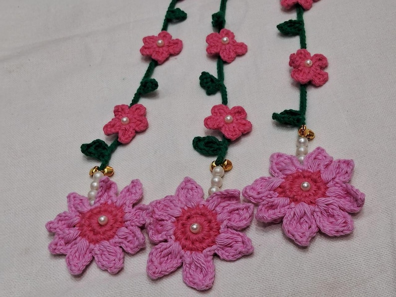 May include: Three crocheted floral garlands featuring dark green stems with small leaves and pink flowers. Each garland terminates in a large, multi-petaled pink flower with a dark pink center, accented by small white beads and gold-toned bells.