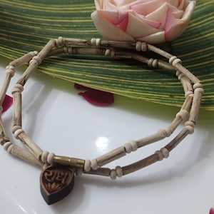 Puede incluir: Un collar artesanal hecho de cuentas y varillas de madera marrón claro. El collar presenta un colgante en forma de corazón marrón oscuro con texto grabado. El collar se exhibe sobre una superficie blanca con una hoja verde y una flor en el fondo.