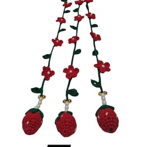 May include: Three crocheted strawberry charms hanging from green vines decorated with small red flowers and white pearl accents. Each strawberry is red with white dots, connected to the vine by a string of white beads and a gold-toned bell. Letter E.