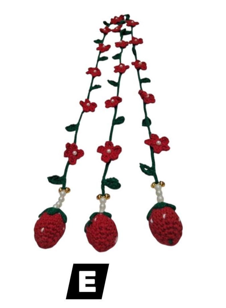 May include: Three crocheted strawberry charms hanging from green vines decorated with small red flowers and white pearl accents. Each strawberry is red with white dots, connected to the vine by a string of white beads and a gold-toned bell. Letter E.