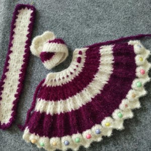 May include: A crocheted baby outfit in white and purple with a matching hat and a long white strap. The outfit is decorated with colorful beads.