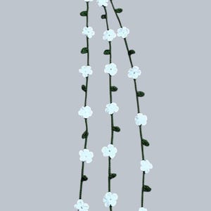 May include: A hanging decoration featuring three dark green crocheted vines suspended from a single dark green ring. Each vine is adorned with small white crocheted flowers and tiny green leaves, ending in a white tassel with small white beads.