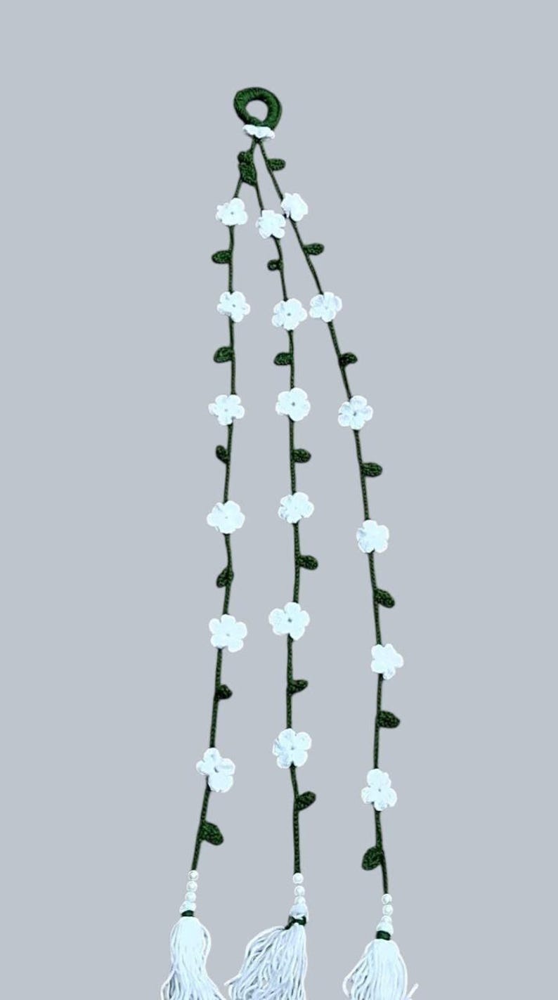 May include: A hanging decoration featuring three dark green crocheted vines suspended from a single dark green ring. Each vine is adorned with small white crocheted flowers and tiny green leaves, ending in a white tassel with small white beads.