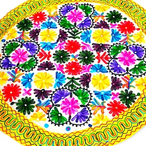 Puede incluir: Un mantel circular blanco con un borde amarillo y un diseño de bordado floral colorido. El bordado presenta una variedad de flores en tonos de rosa, rojo, morado, azul, verde y amarillo.