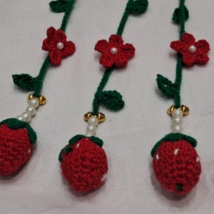 May include: Three crocheted strawberry ornaments hanging from green yarn stems. Each stem features a red crocheted flower with a central white bead, green leaves, a string of white pearls, and a gold-toned bell above the red strawberry fruit.
