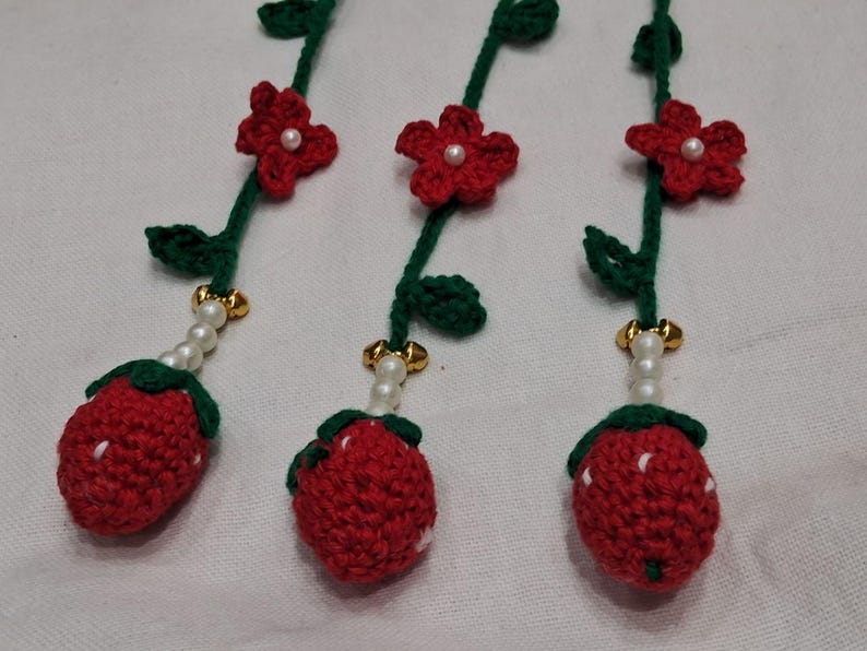 May include: Three crocheted strawberry ornaments hanging from green yarn stems. Each stem features a red crocheted flower with a central white bead, green leaves, a string of white pearls, and a gold-toned bell above the red strawberry fruit.