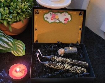 May include: A black box with a gold lid and a floral design. The box contains three black candles with gold glitter, a small glass jar with a black substance, and a black and gold star charm. The box is labeled "Carla & Jordan Magical Boutique".