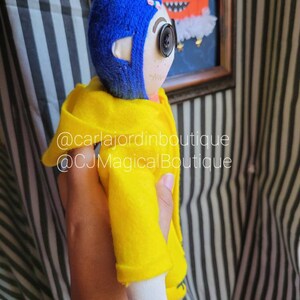 May include: A handmade doll wearing a yellow raincoat and a blue yarn hat. The doll has a button eye and a stitched smile. The doll is holding a small pumpkin in its hand. The doll is standing in front of a black and white striped background. @carlajordinboxboutique @CJMagicalBoutique