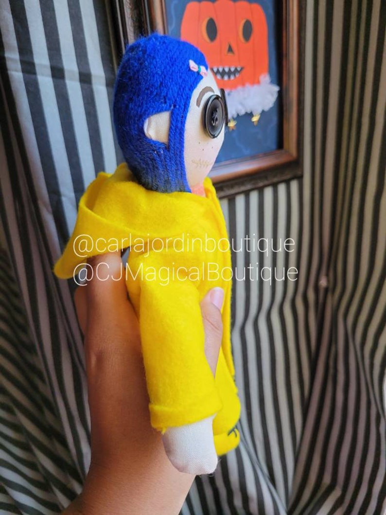 May include: A handmade doll wearing a yellow raincoat and a blue yarn hat. The doll has a button eye and a stitched smile. The doll is holding a small pumpkin in its hand. The doll is standing in front of a black and white striped background. @carlajordinboxboutique @CJMagicalBoutique