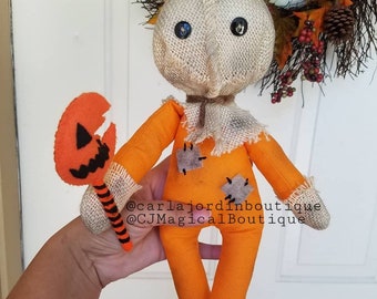 May include: A handmade orange and brown fabric doll with a burlap head and black eyes. The doll is holding a felt orange pumpkin with a black jack-o-lantern face. The doll is standing on a hand. The text "@carlajordanboutique @CJMagicalBoutique" is visible on the doll's body.