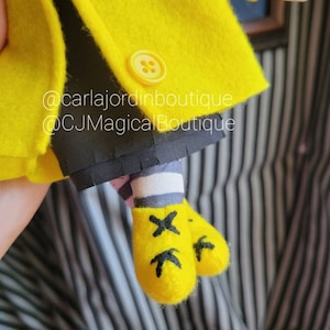 May include: A yellow felt doll wearing a yellow coat with black buttons and black and white striped socks. The doll is wearing yellow felt shoes with black X markings. The doll is holding a white felt object. The image includes the text "@carlajordinboutique @CJMagicalBoutique".