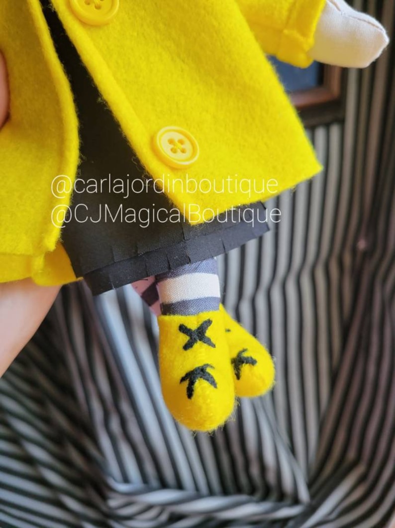 May include: A yellow felt doll wearing a yellow coat with black buttons and black and white striped socks. The doll is wearing yellow felt shoes with black X markings. The doll is holding a white felt object. The image includes the text "@carlajordinboutique @CJMagicalBoutique".