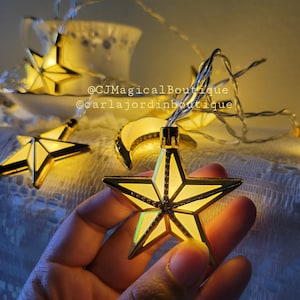 May include: A string of fairy lights with gold star and crescent moon shaped bulbs. The lights are strung on a clear wire and are lit up. The lights are on a white lace tablecloth. The text '@CJMagicalBoutique @carlajordinboutique' is visible in the image.